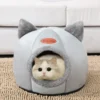 A cute cat peeking out from the entrance of its grey cat cave bed, with cozy ear accents on top, looking relaxed and at home.